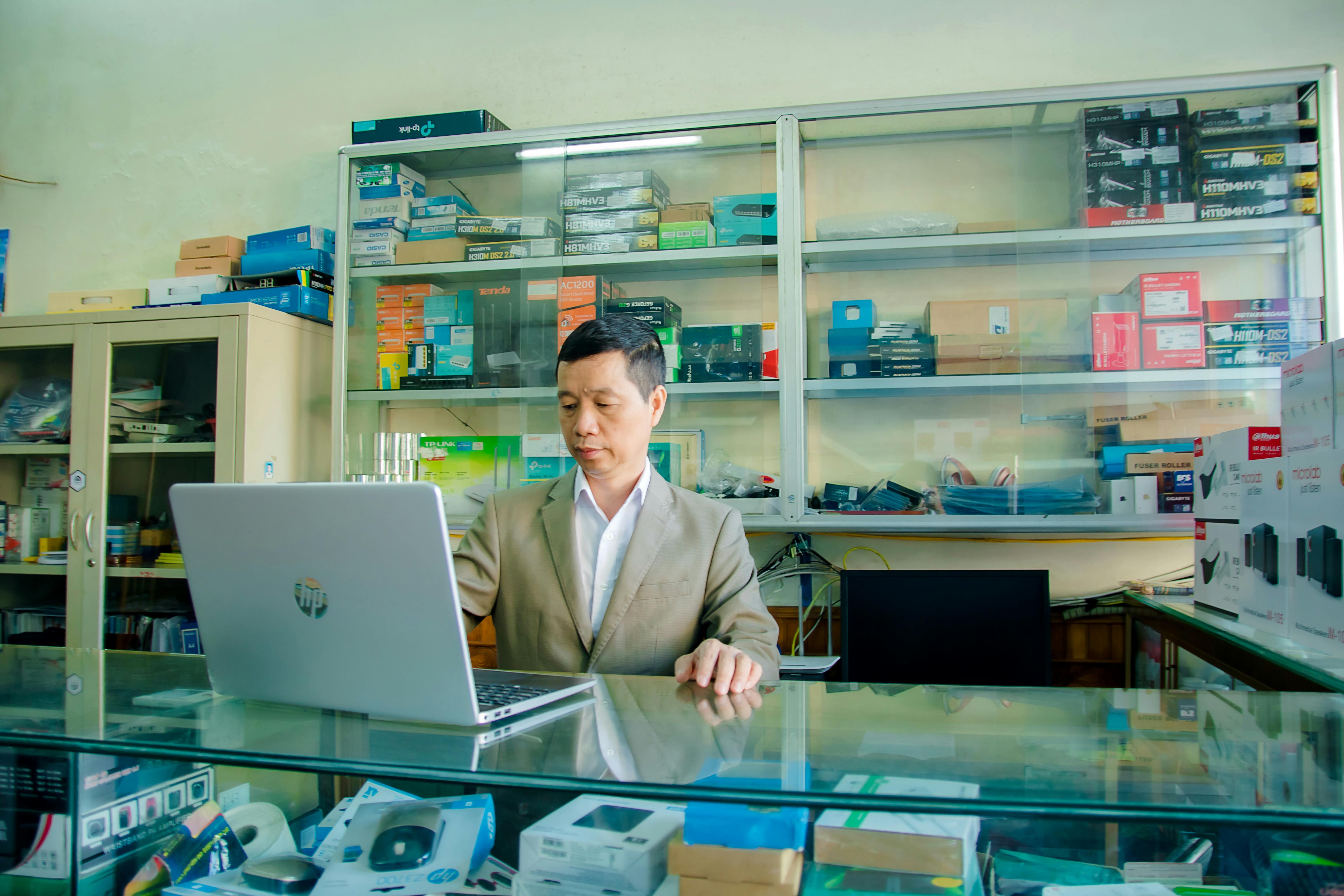 A man in a beige suit is working on a laptop at a glass counter. Behind him, shelves display various electronic items and boxes.