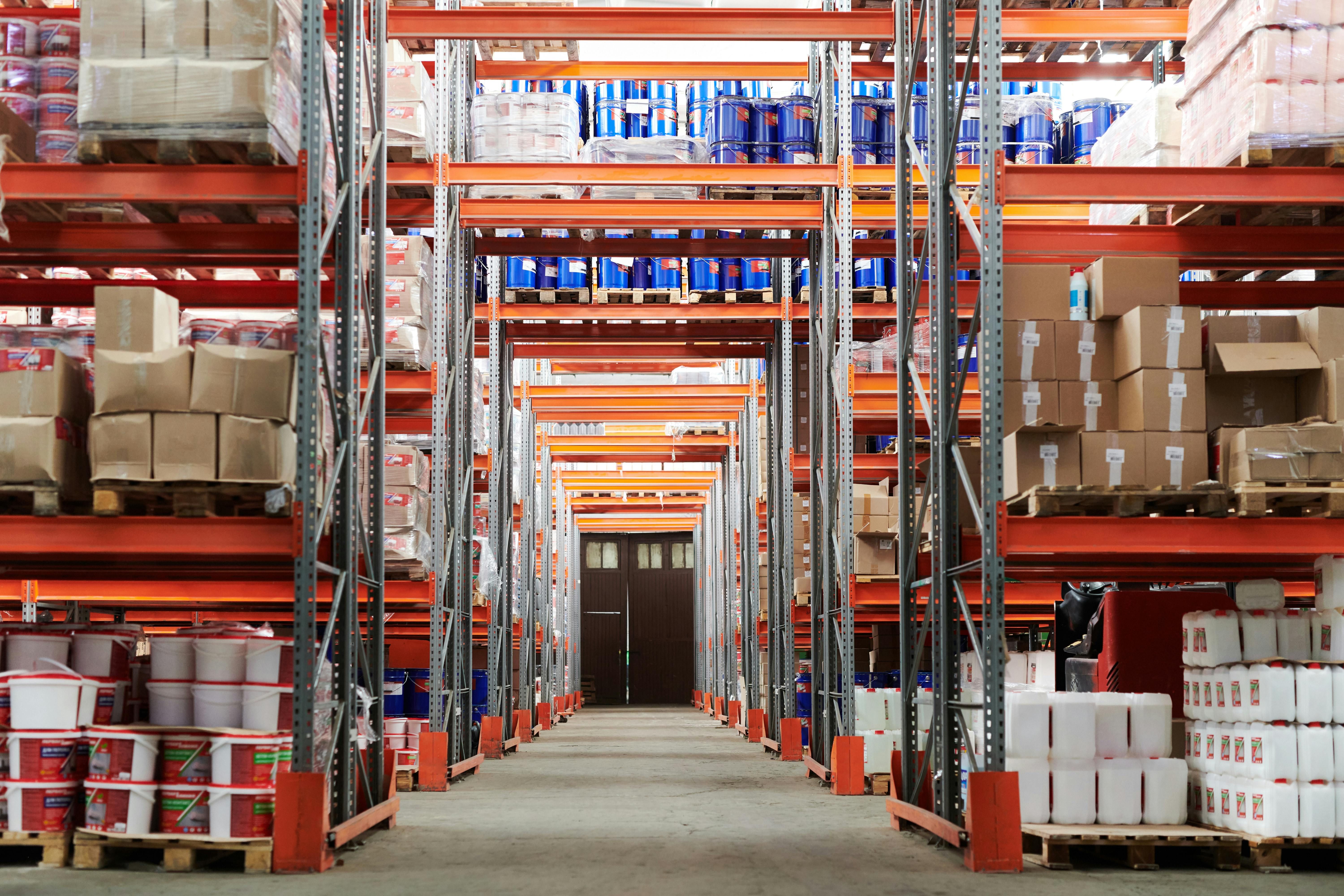 A warehouse interior featuring tall shelves filled with boxes, containers, and assorted items. A central aisle runs through the space, ending at a closed door in the background.