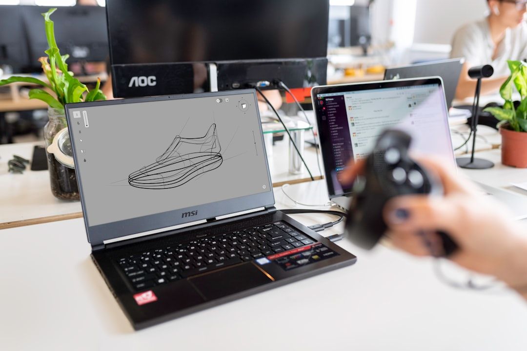 A workspace featuring a laptop showing a shoe design, a person using a 3D mouse, and another laptop open to a messaging app.